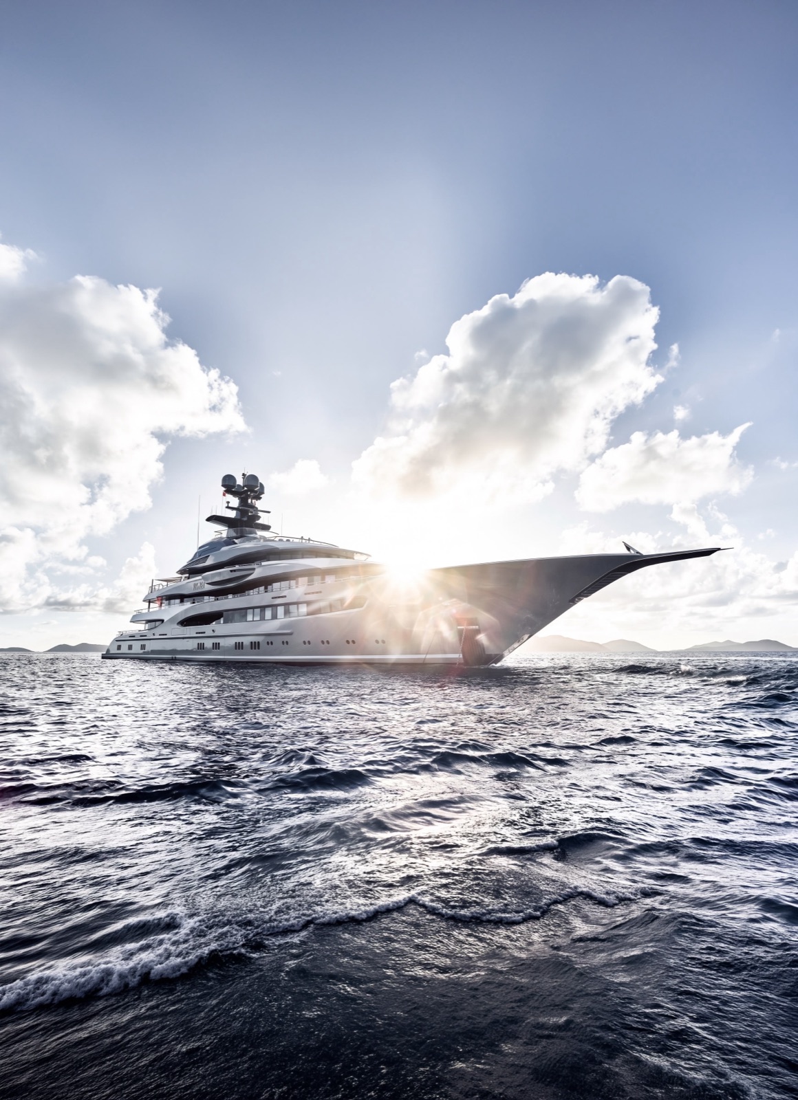 A large white motor yacht under way on open water, backlit by the sun with cloud building behind her.