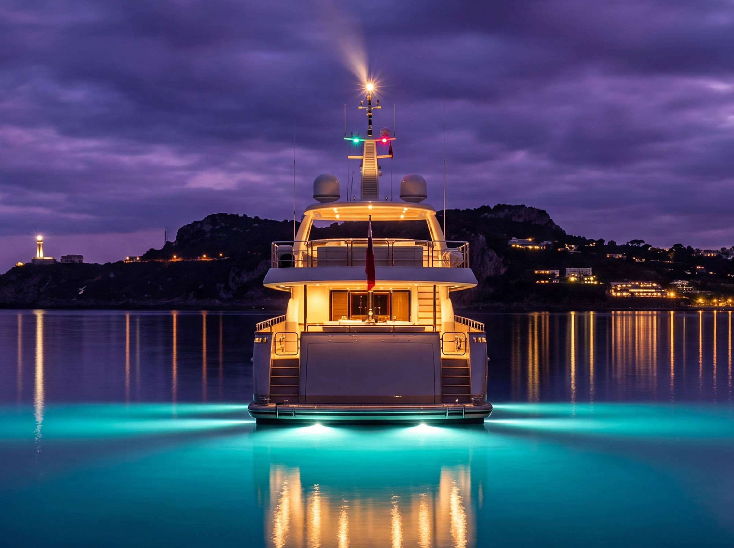 A motor yacht lit from within at dusk, her underwater lights glowing off a calm harbour