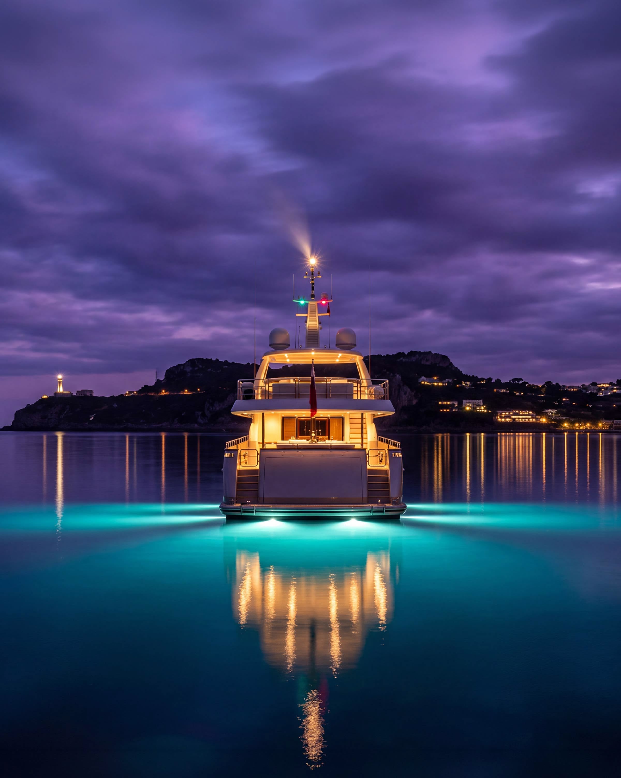 A motor yacht lit up at dusk off a headland, her lights reflected in turquoise water under a purple sky.