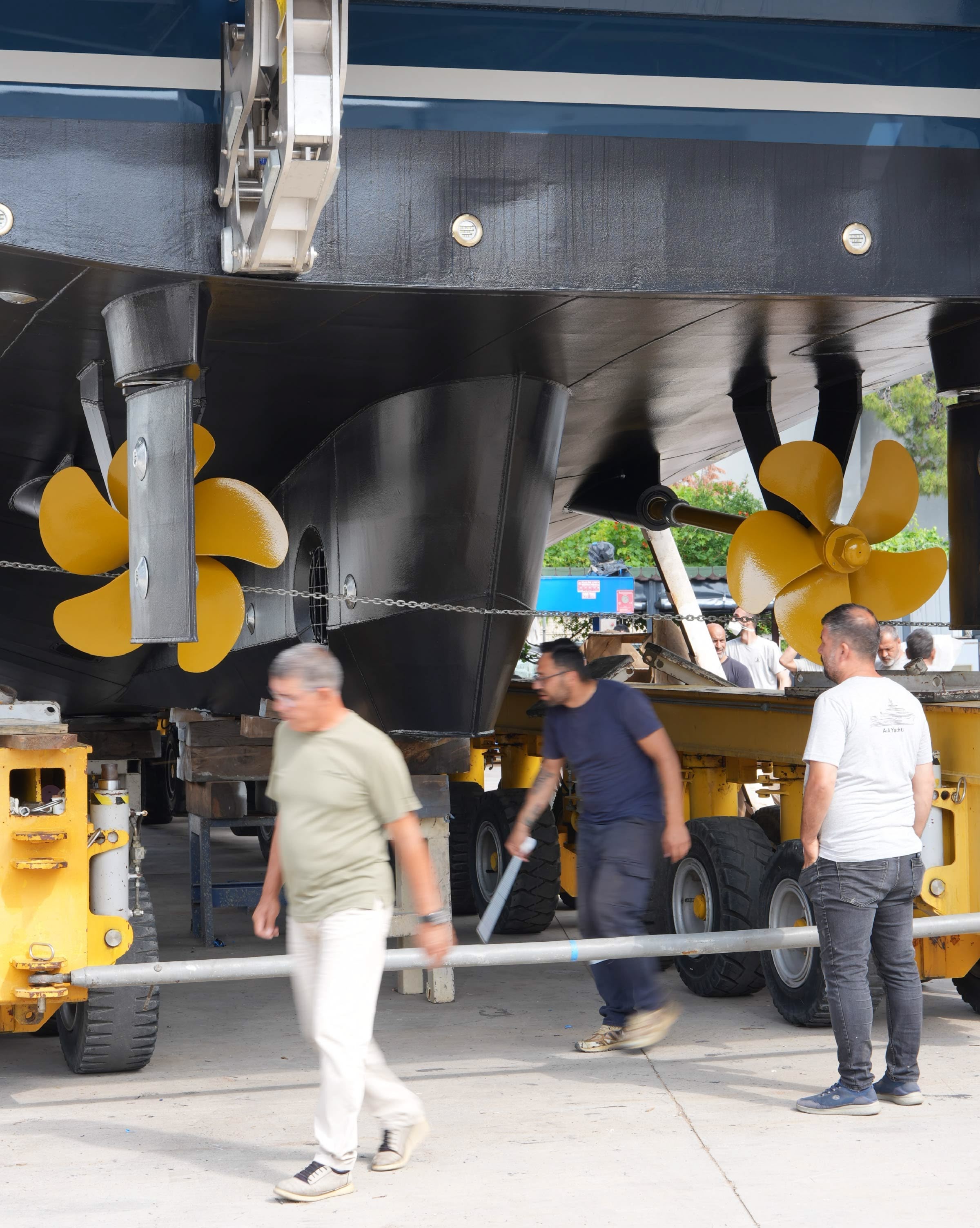 A yacht held in a travel hoist out of the water, her gold propellers and shafts exposed while the yard team looks underneath.